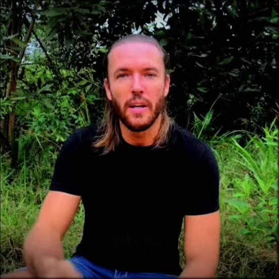 A bearded man with long hair seated outdoors in dense greenery, speaking calmly to the camera